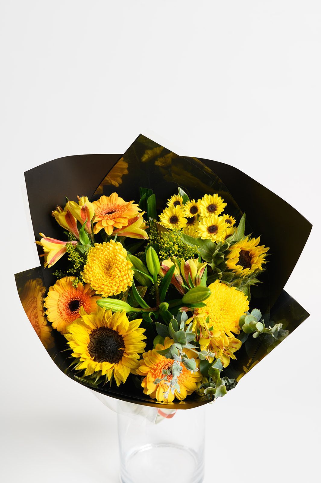 Bouquet of yellow and orange sunflowers wrapped in dark paper, shown in a clear vase.