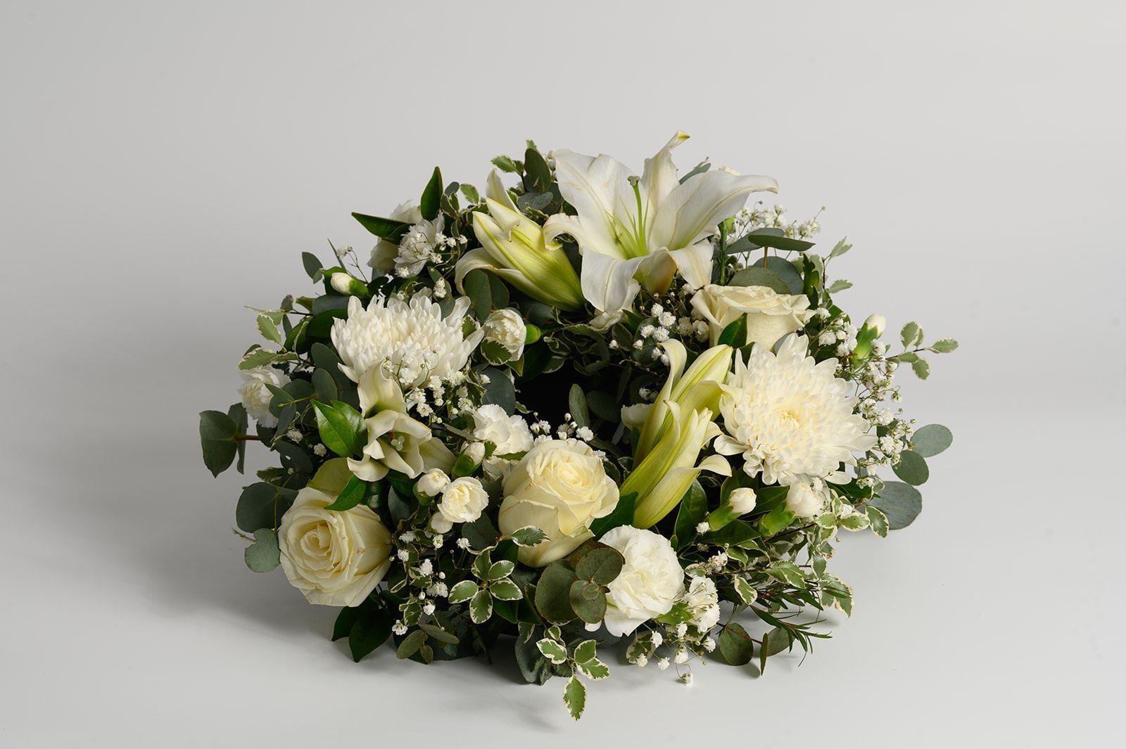 White floral wreath with lilies, roses, and greenery on a light gray background
