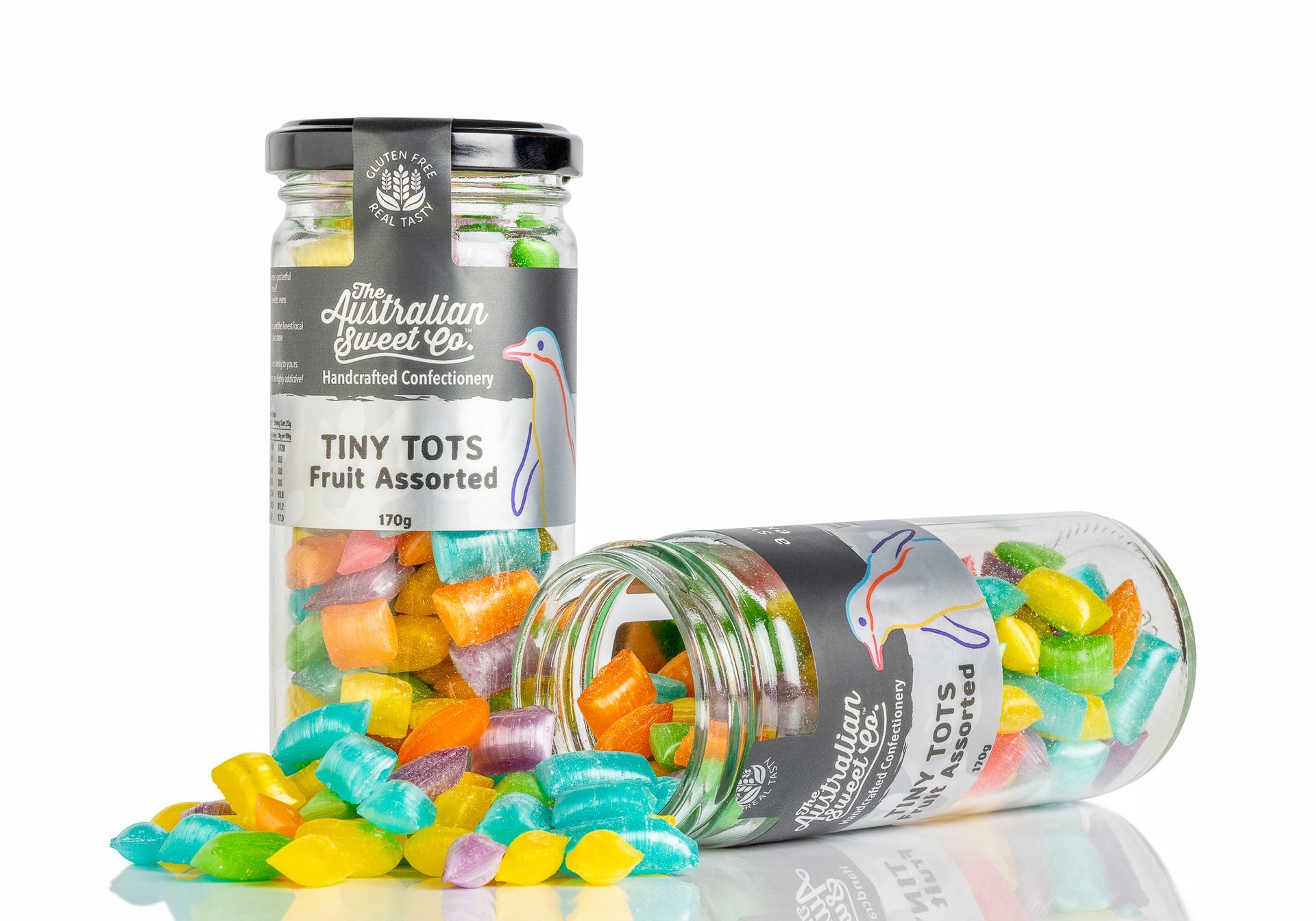 Two glass jars of colorful fruit-shaped candy, one upright and one tipped over, spilling candies on white background