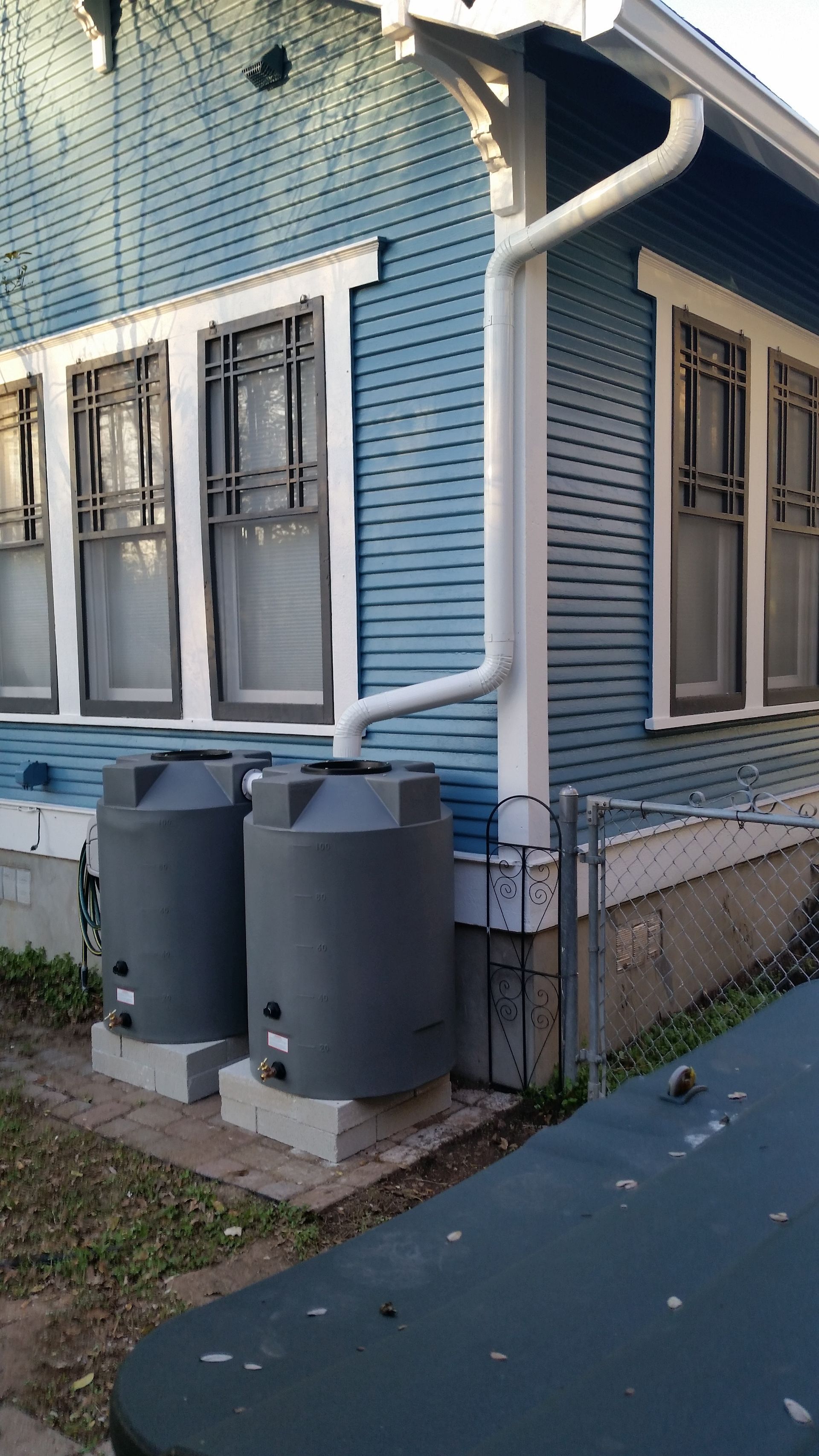 Two gray rain barrels beside a blue house, connected to a white gutter.