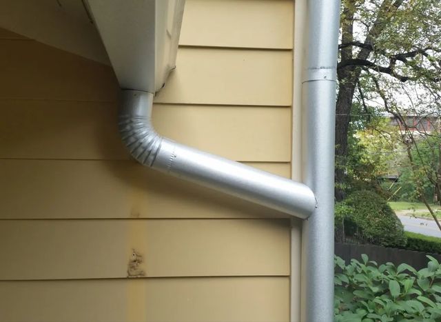 Galvanized metal rain gutter and downspout on a white house with patterned siding.