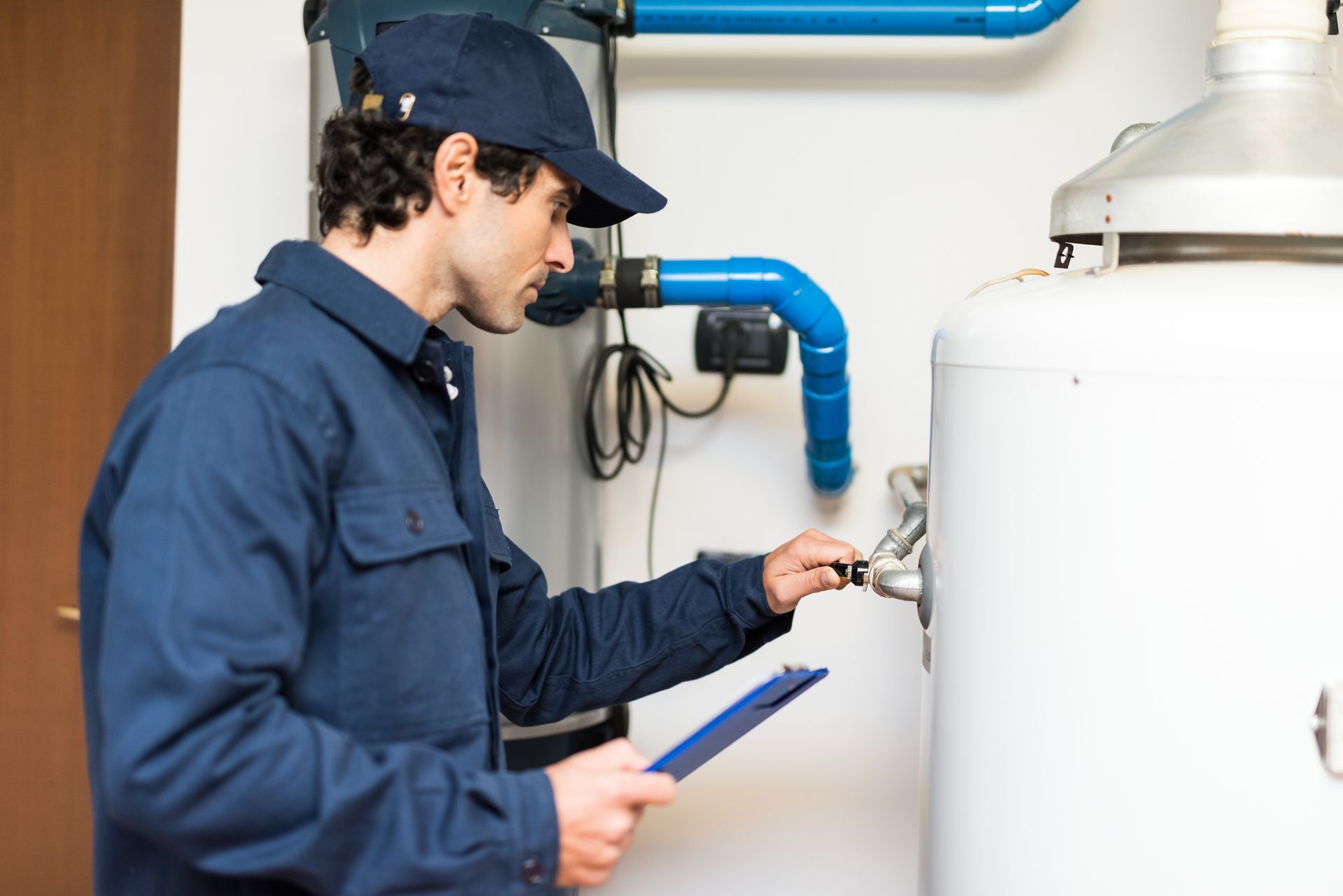 A person in a blue uniform and cap inspects a white water heater while holding a clipboard. A person in a blue uniform and cap inspects a white water heater while holding a clipboard.