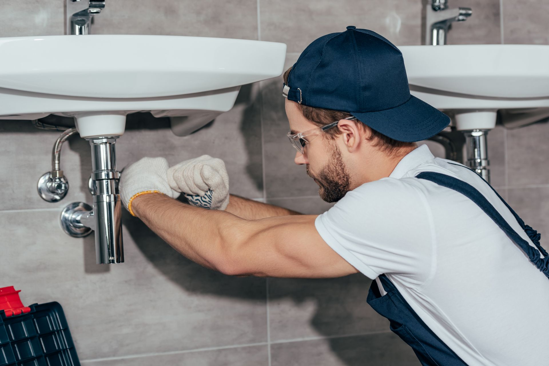 Plumber in safety glasses and gloves tightening a chrome drain trap beneath a white bathroom sink.