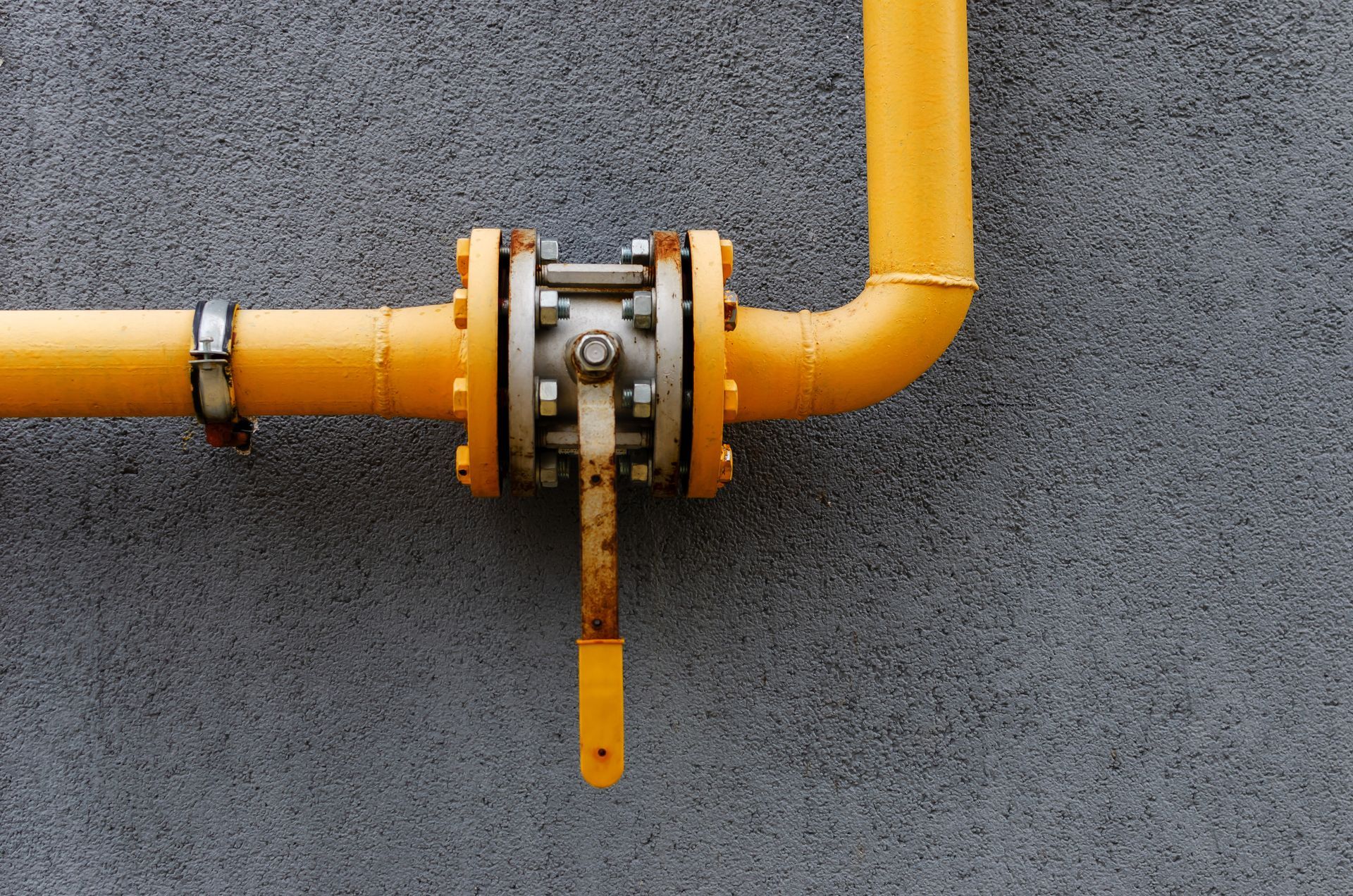 A yellow pipe mounted on a gray textured wall features a large, flanged ball valve with a vertical handle. A yellow pipe mounted on a gray textured wall features a large, flanged ball valve with a vertical handle.