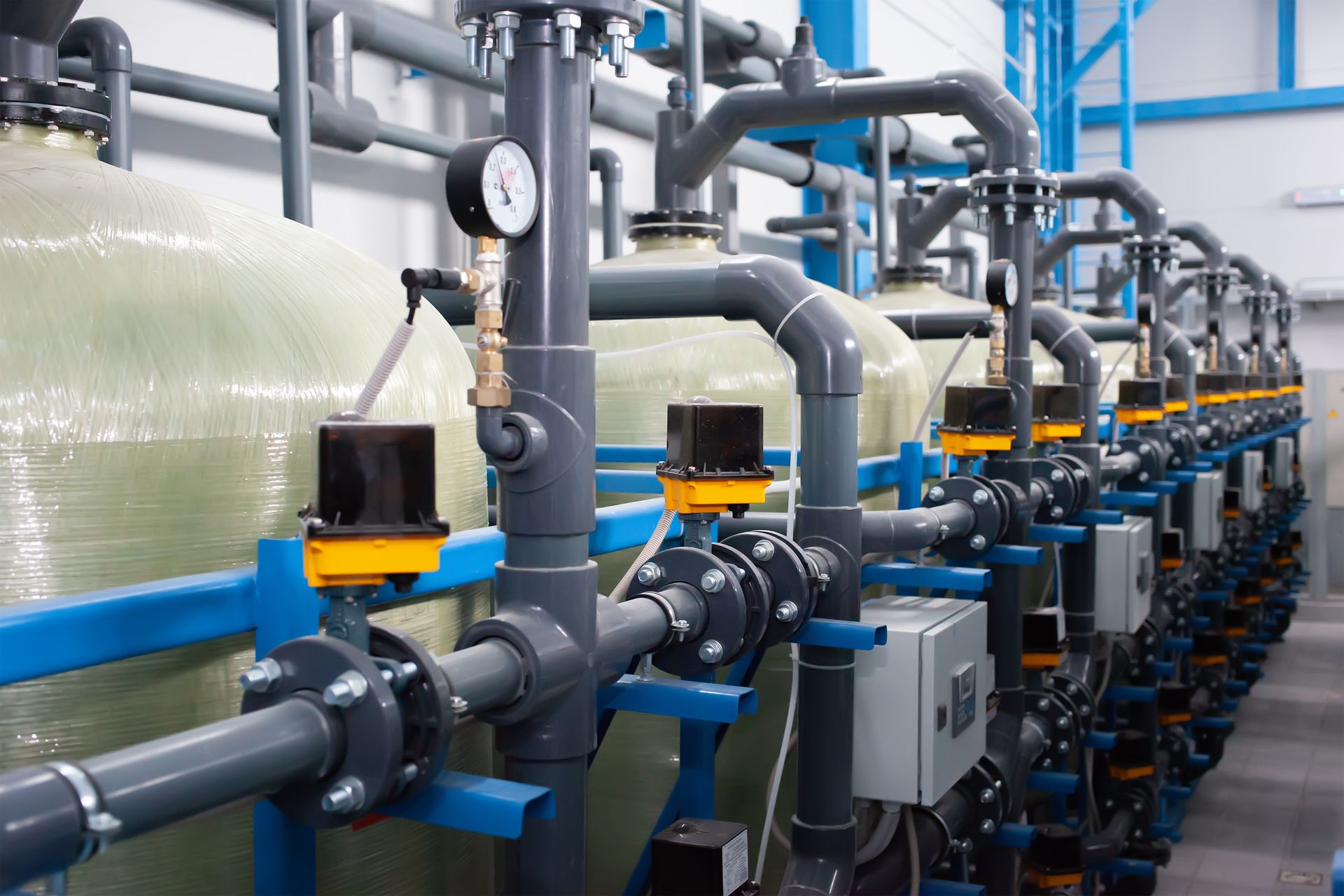 Industrial water treatment facility featuring a row of large beige storage tanks connected by grey pipes and valves. Industrial water treatment facility featuring a row of large beige storage tanks connected by grey pipes and valves.