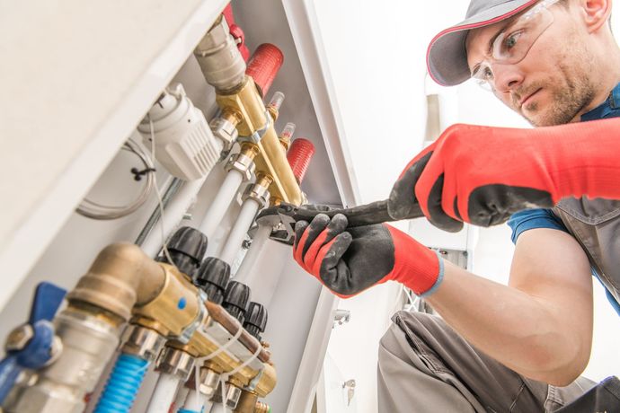 A technician in safety gear uses a wrench to adjust a wall-mounted heating system manifold with copper and white pipes. A technician in safety gear uses a wrench to adjust a wall-mounted heating system manifold with copper and white pipes.
