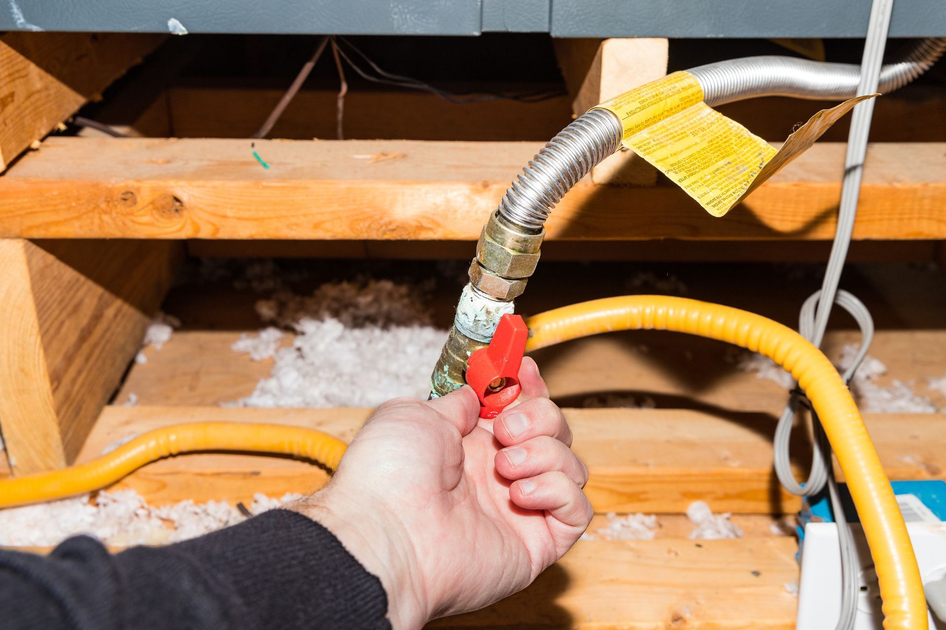 A hand turns the red lever on a gas shut-off valve connected to a flexible metal supply line in an attic. A hand turns the red lever on a gas shut-off valve connected to a flexible metal supply line in an attic.