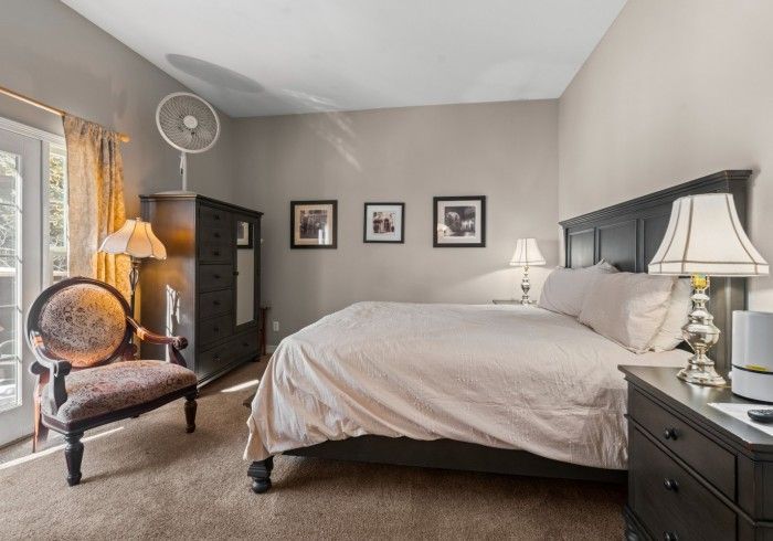 A bedroom with a bed , chair , nightstand , lamp and fan.