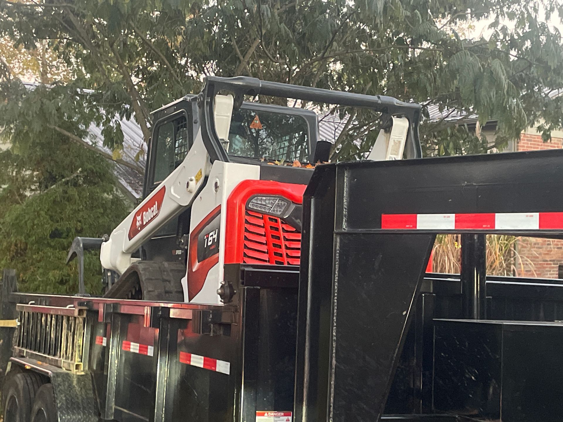 A white and red Bobcat skid-steer loader sits loaded onto a black flatbed trailer outdoors.