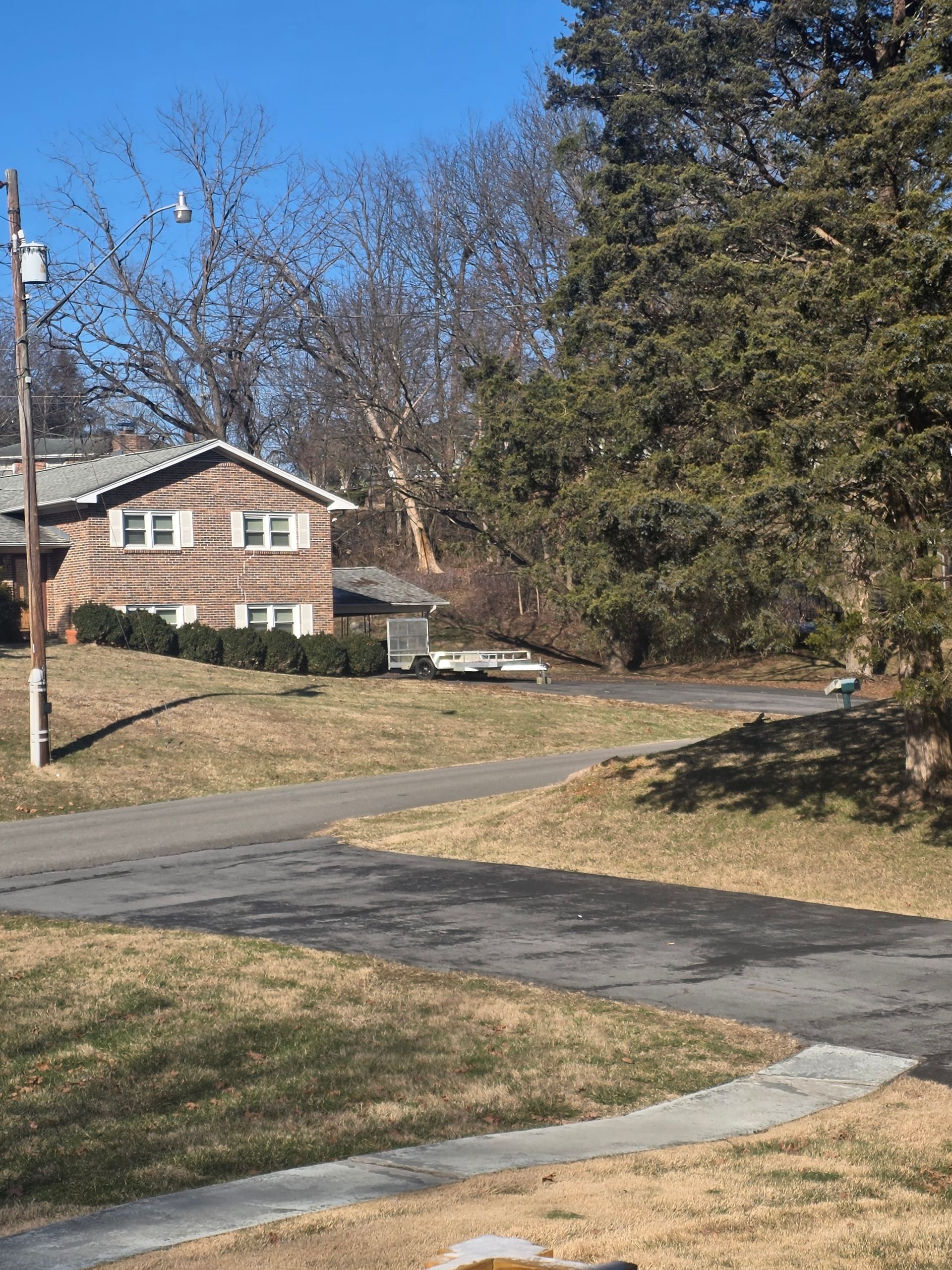 A two-story brick house with white shutters sits at the end of a paved driveway, surrounded by trees under a clear blue sky.