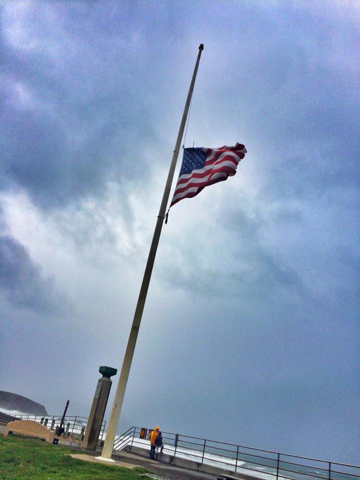 American flag at half-staff on a flagpole near a coastline, under a cloudy sky.