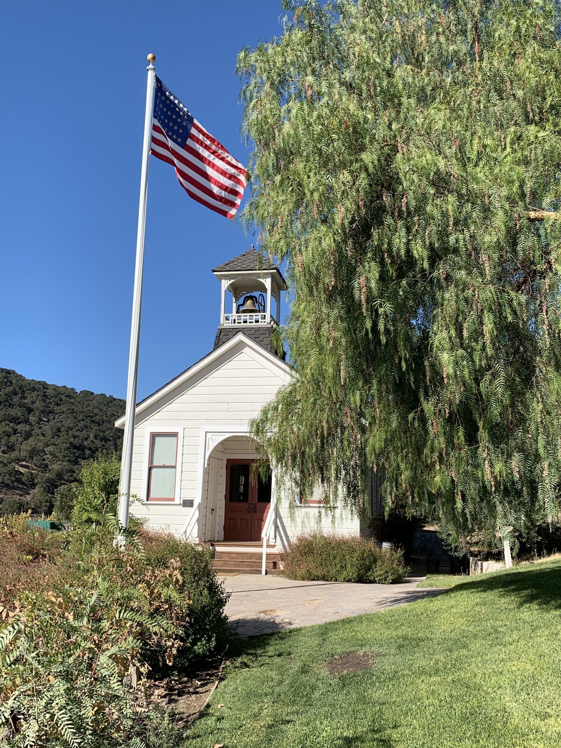 Small white schoolhouse with American flag waving. Bell tower, red door, green bushes, and a pathway. Sunny day.