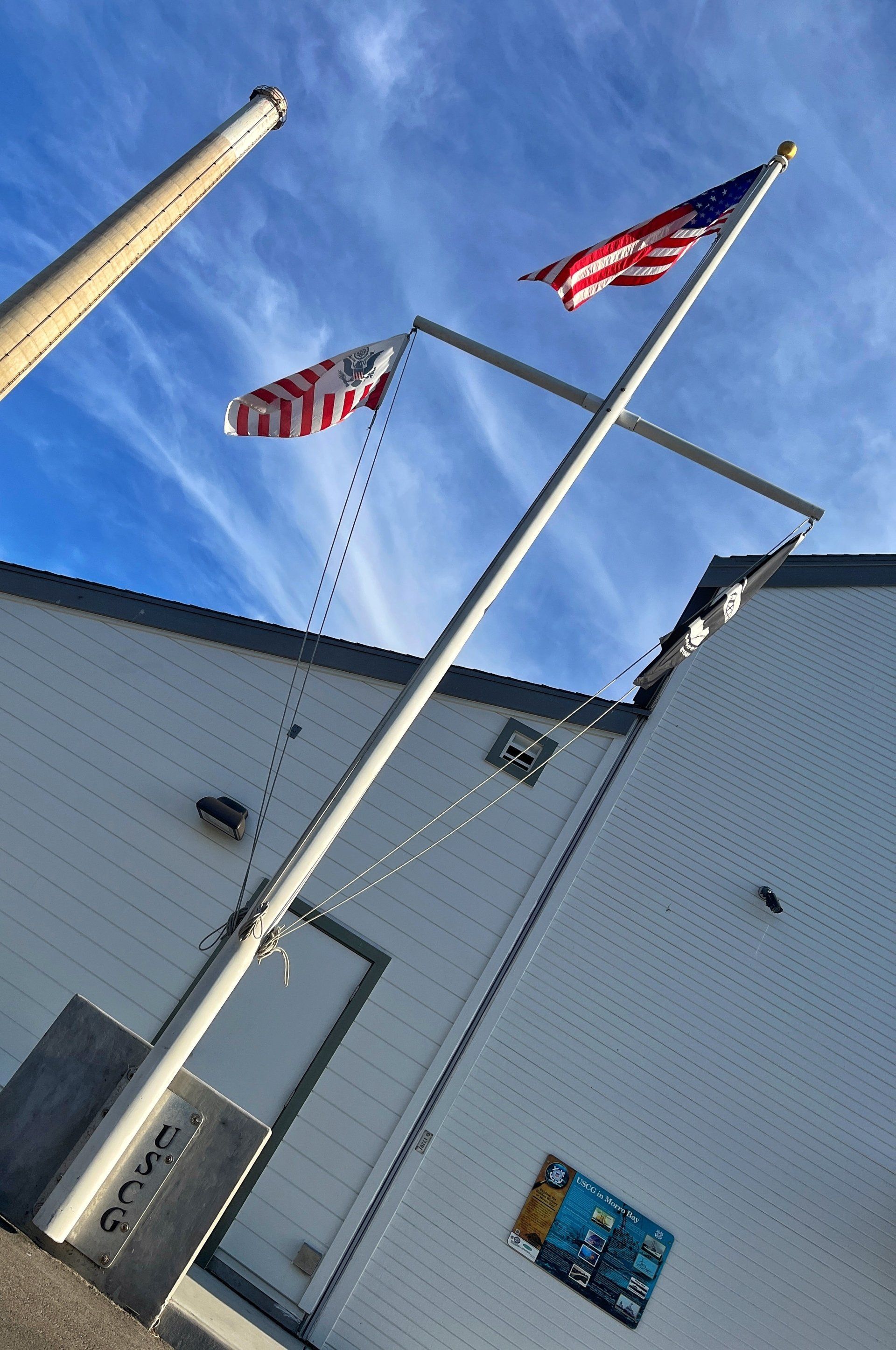 Two flags waving in the wind atop a white building: US flag and a second, red and white striped flag. Blue sky.