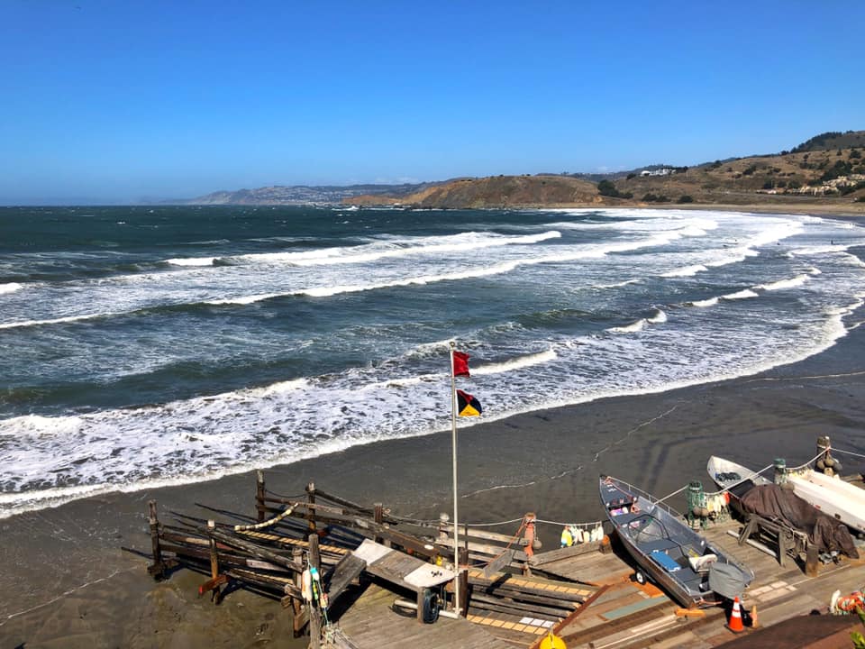 View of a beach with waves, a flag, boats, and a hillside under a blue sky.