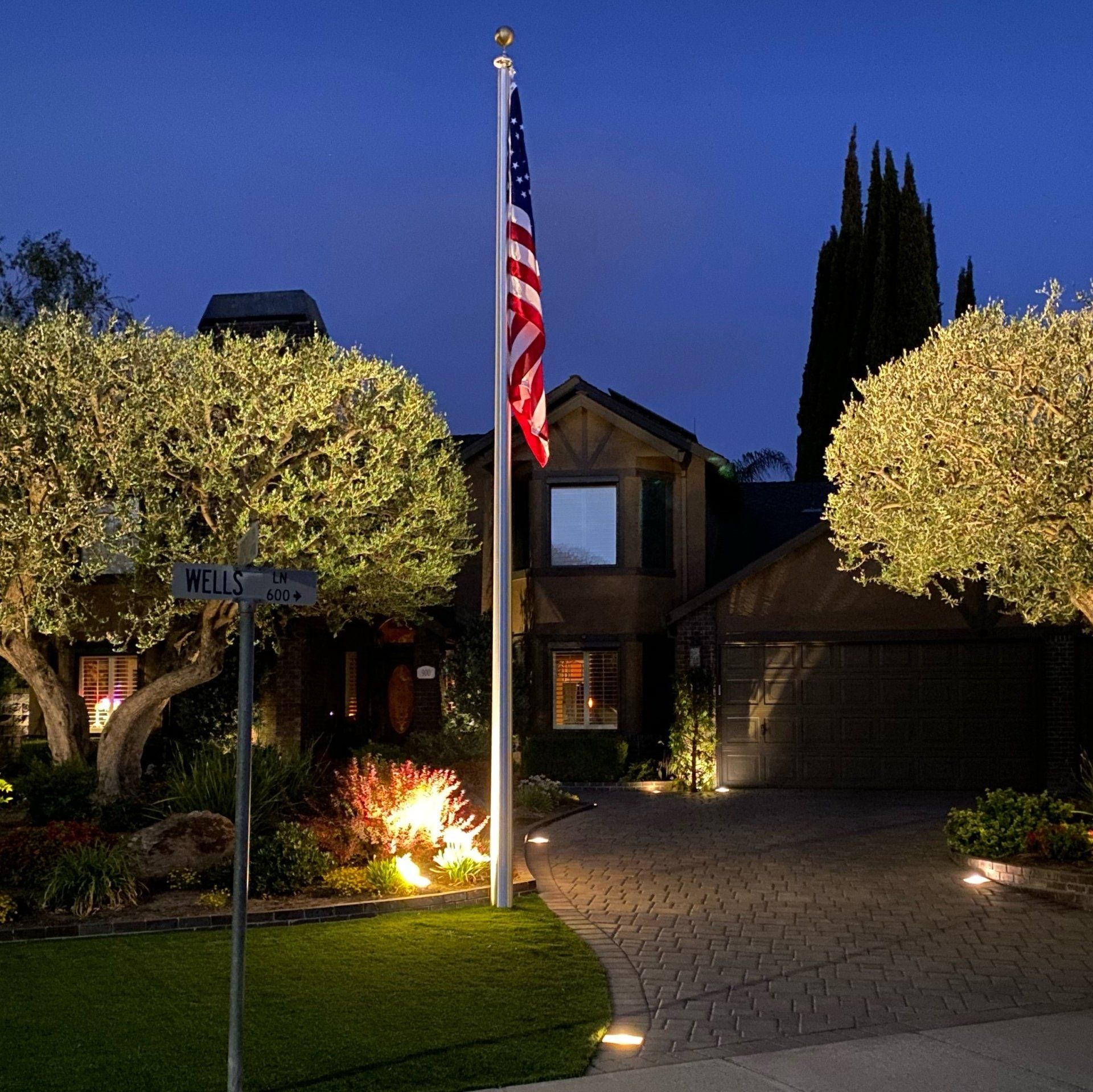 A flag flies on a pole in front of a house, lit by ground lights, at dusk.
