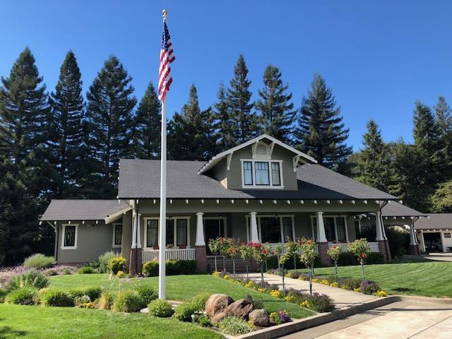 A Craftsman-style house with an American flag, surrounded by a green lawn and tall trees under a blue sky.
