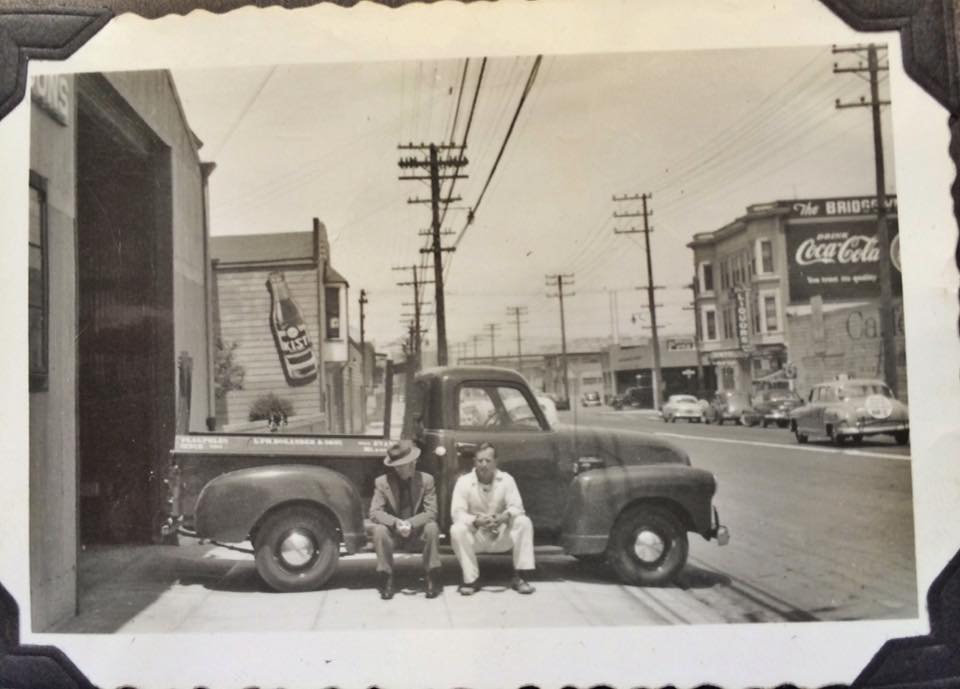 Two men sit on a pickup truck. A Coca-Cola sign and buildings are visible on the street.
