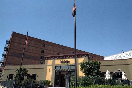 Restaurant building with flags. Brown brick building in the background, tan building in the foreground with outdoor seating.