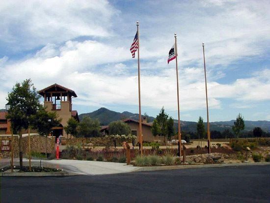 Entrance to a building with American and California flags, trees, and mountains under a cloudy sky.