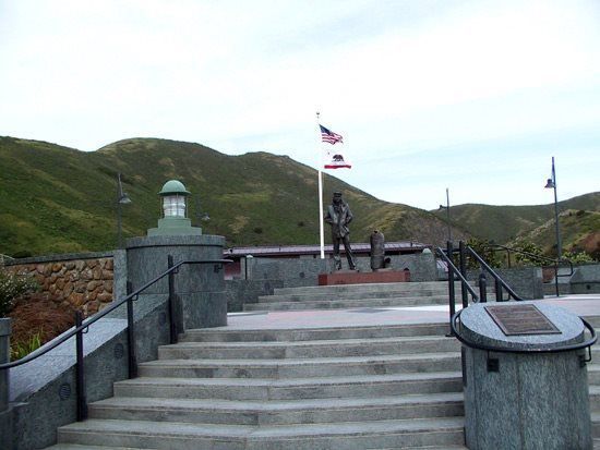Statue on steps with US and California flags, hills in background.