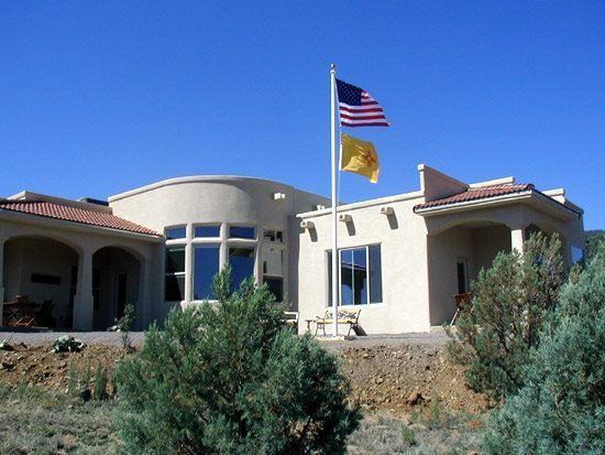 House with beige stucco walls, red tile roof, and US and New Mexico flags on a pole.