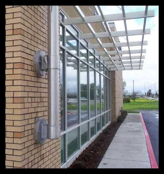 Brick building with large windows, metal awning, and exhaust pipe. Concrete sidewalk along the windows.