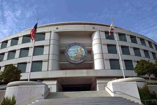 Circular, beige state government building with US and California flags, under blue sky.