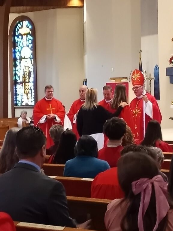 Clergy in red robes stand at the altar during a church ceremony, viewed from the pews.