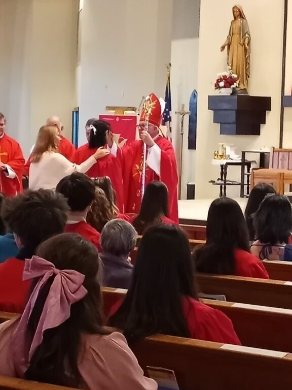 Clergy in red vestments lead a church procession past seated worshippers and a statue on the wall.