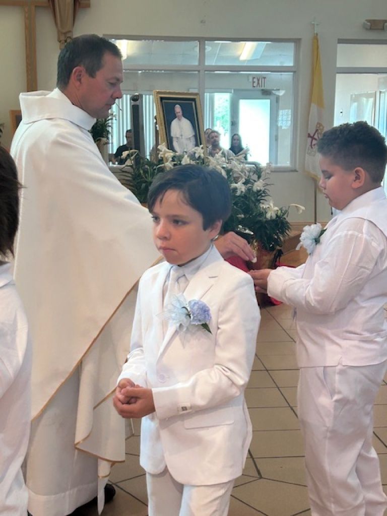People in white suits gathered in a bright church near floral decorations and a statue.