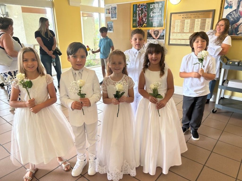 Children in white outfits holding flowers in a bright hallway, posing for a group photo