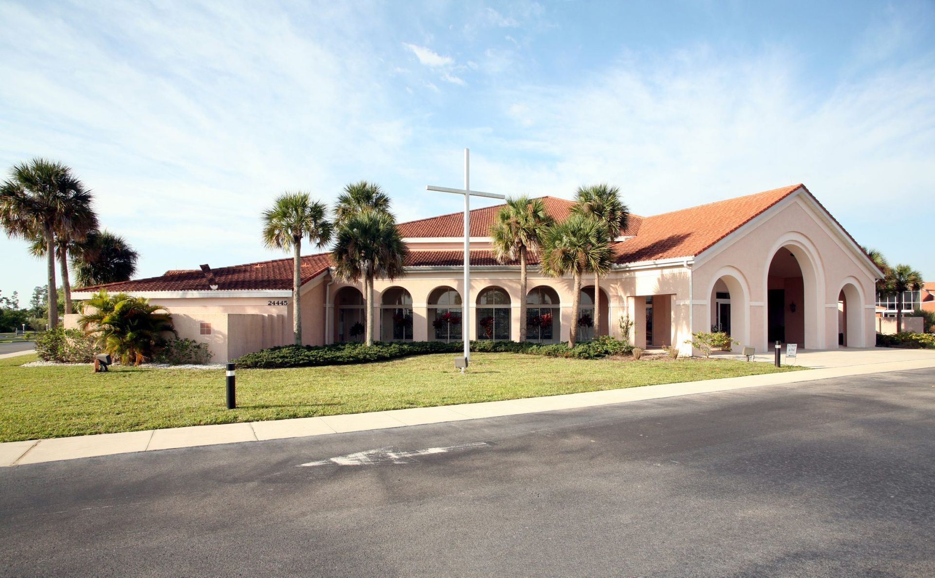 Spanish-style church with red tile roof, arched entrance, and palm trees beside a parking lot