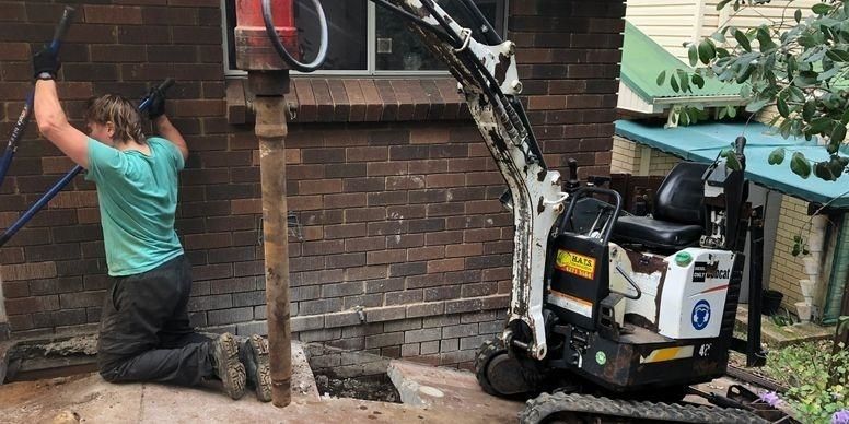 A Man Is Kneeling In Front Of A Brick Wall Next To A Small Excavator — Affordable Underpinning Solutions In Shell Cove, NSW