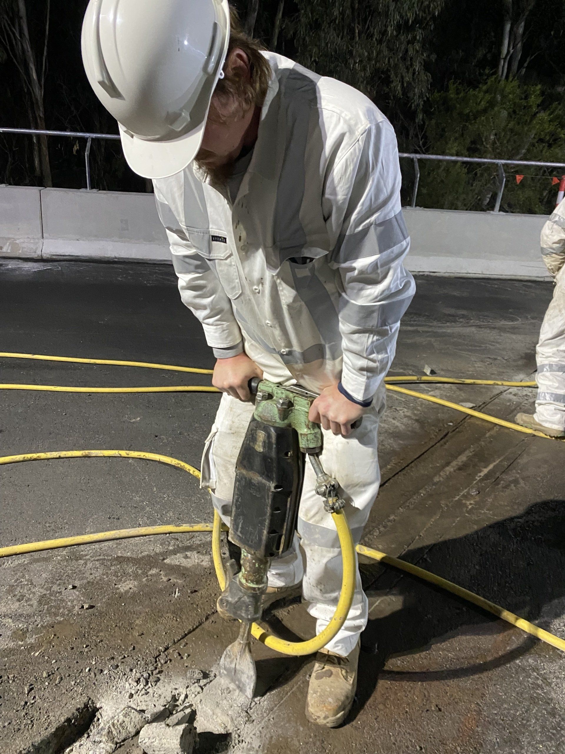 Worker in white hard hat using a jackhammer on a cracked road surface outdoors.