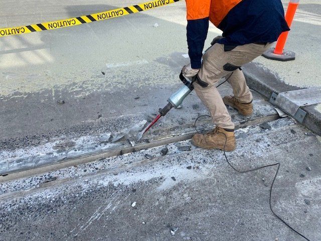 Worker using a jackhammer to break concrete at a road repair site, with caution tape and cones nearby
