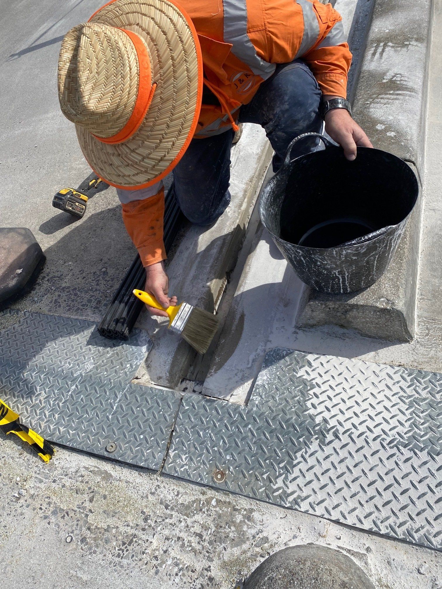 Worker in orange jacket sealing a pavement crack with black tar from a bucket.