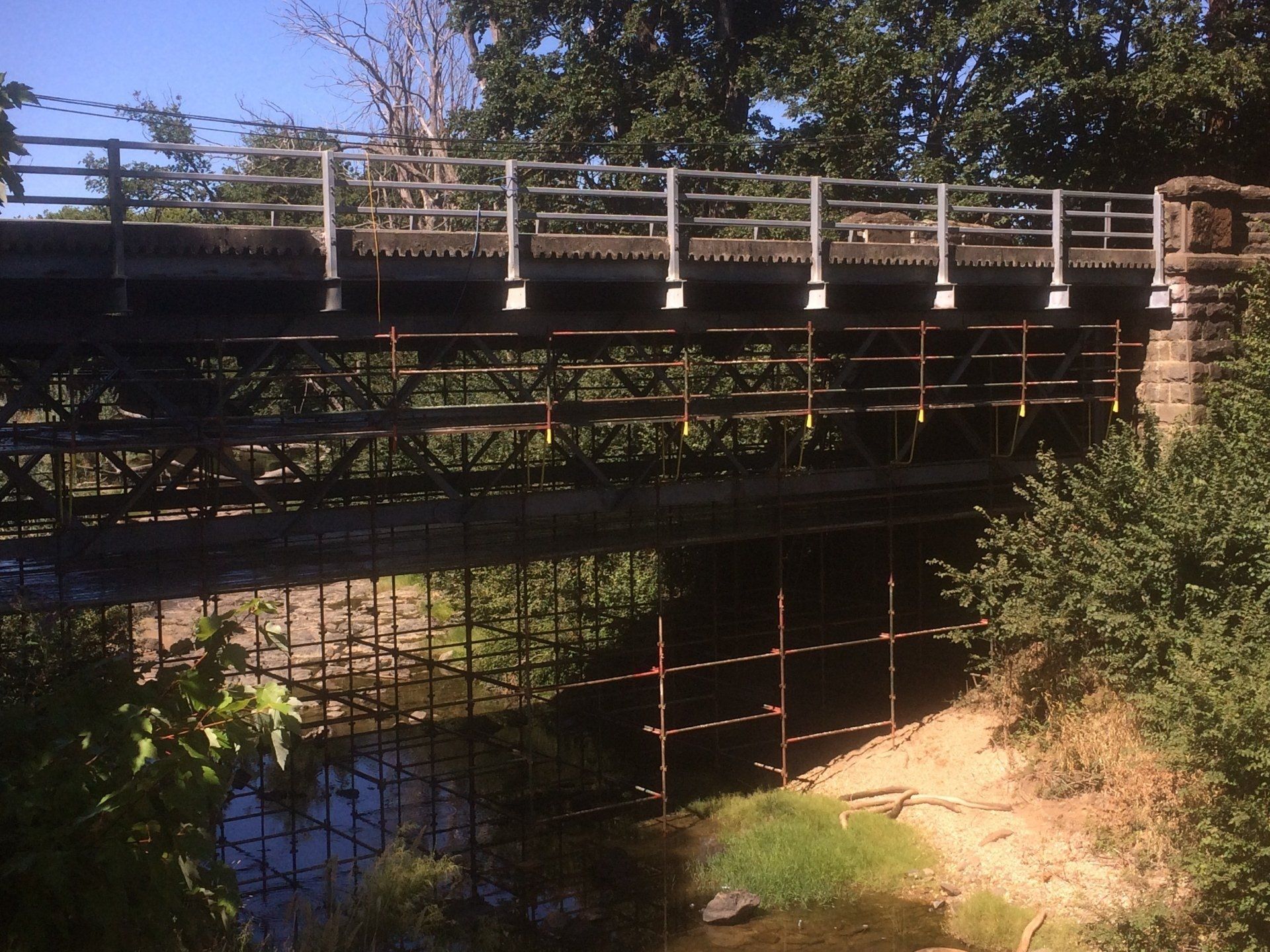 Wooden bridge over a narrow creek, with metal railings and trees in bright sunlight