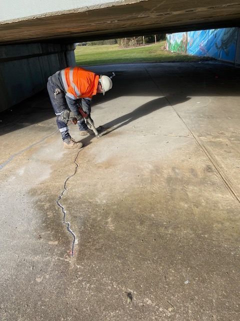 Worker in orange vest repairing a cracked concrete path under a bridge.