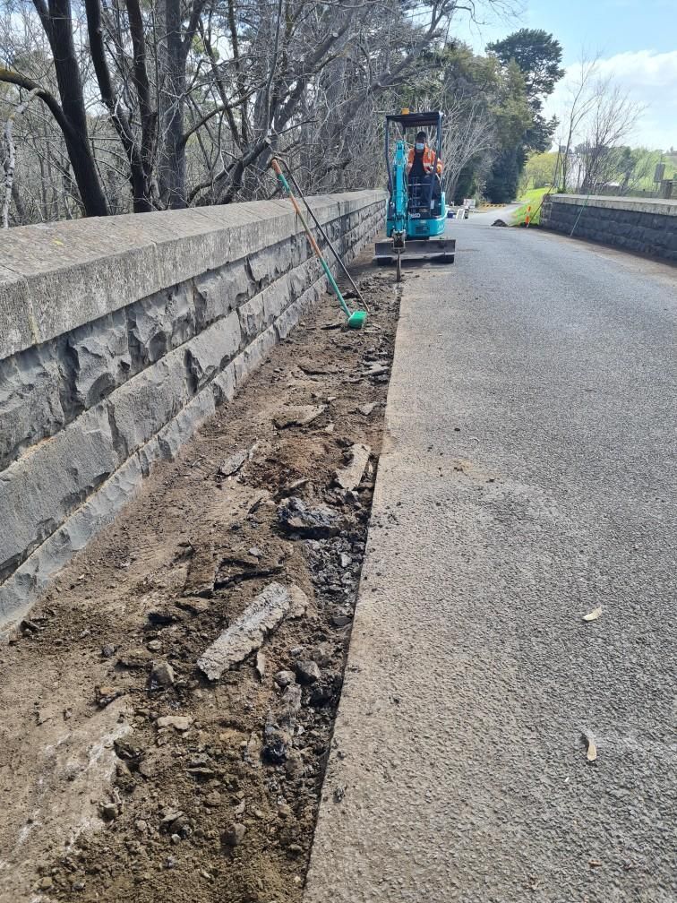 Roadside trench beside stone wall, with a small blue construction machine on the pavement.