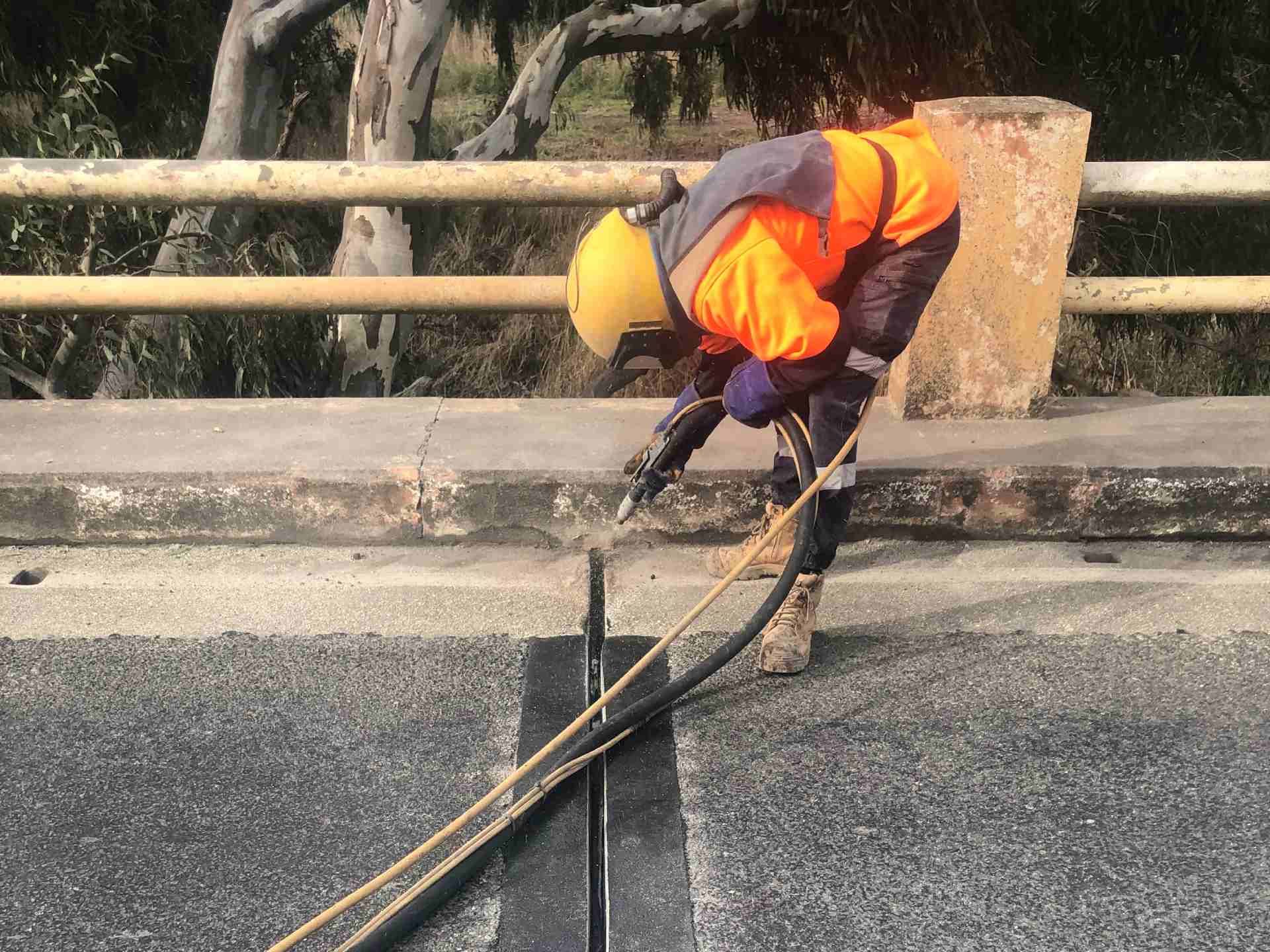 Worker in orange safety gear applying material to a road surface with a hose and tool