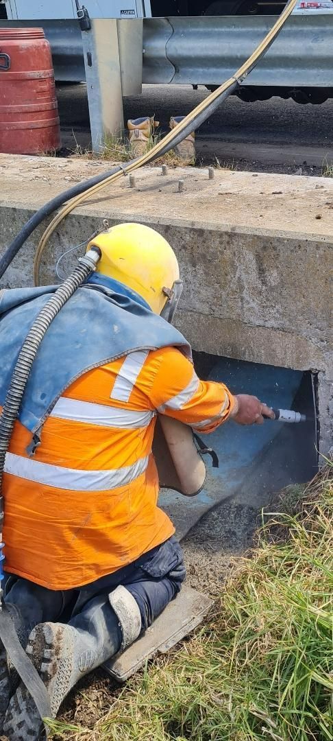 Worker in orange safety vest kneels by a concrete culvert, inspecting the opening under a road.