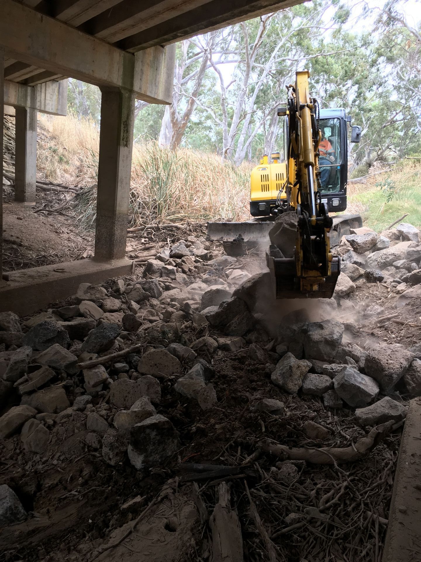 Yellow excavator clearing rocks under a concrete overpass in a debris-filled creek bed