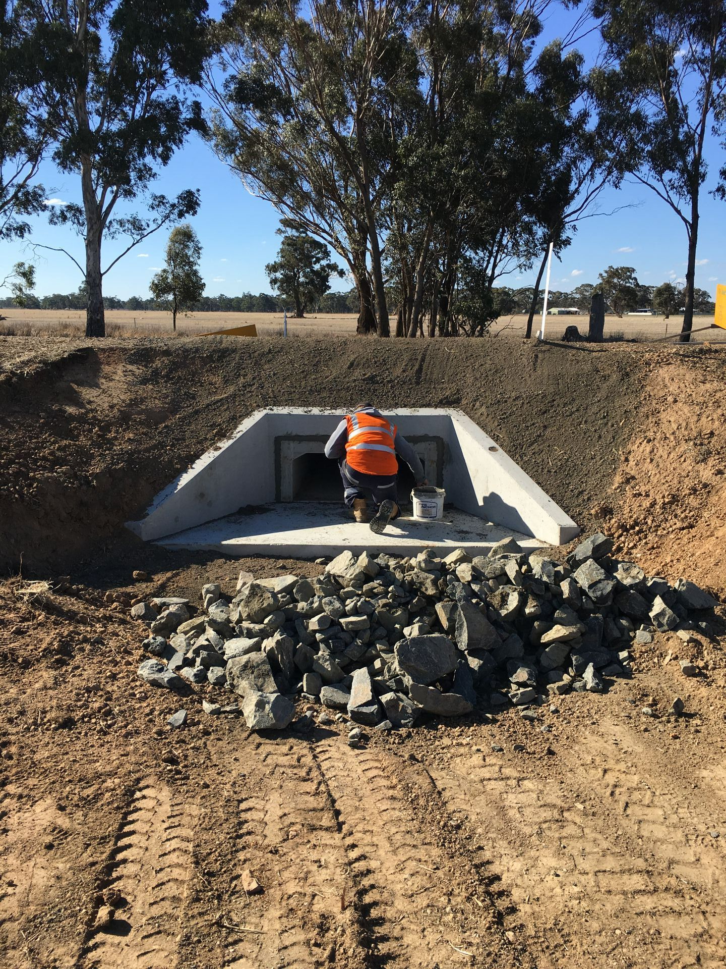 Construction worker in orange vest near a concrete drainage culvert, with rocks and dirt in a wooded field