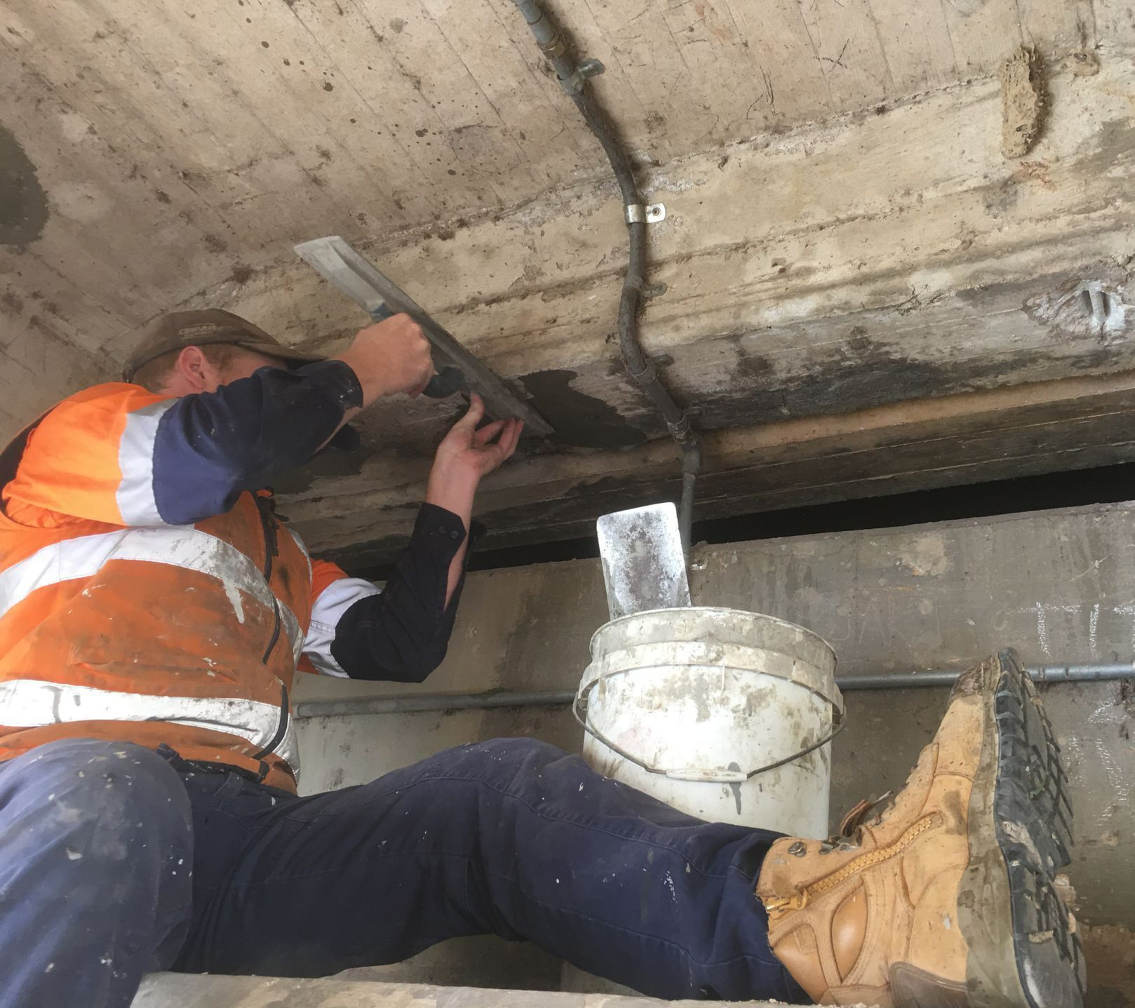 Worker in orange safety vest chiseling concrete overhead inside a narrow crawl space