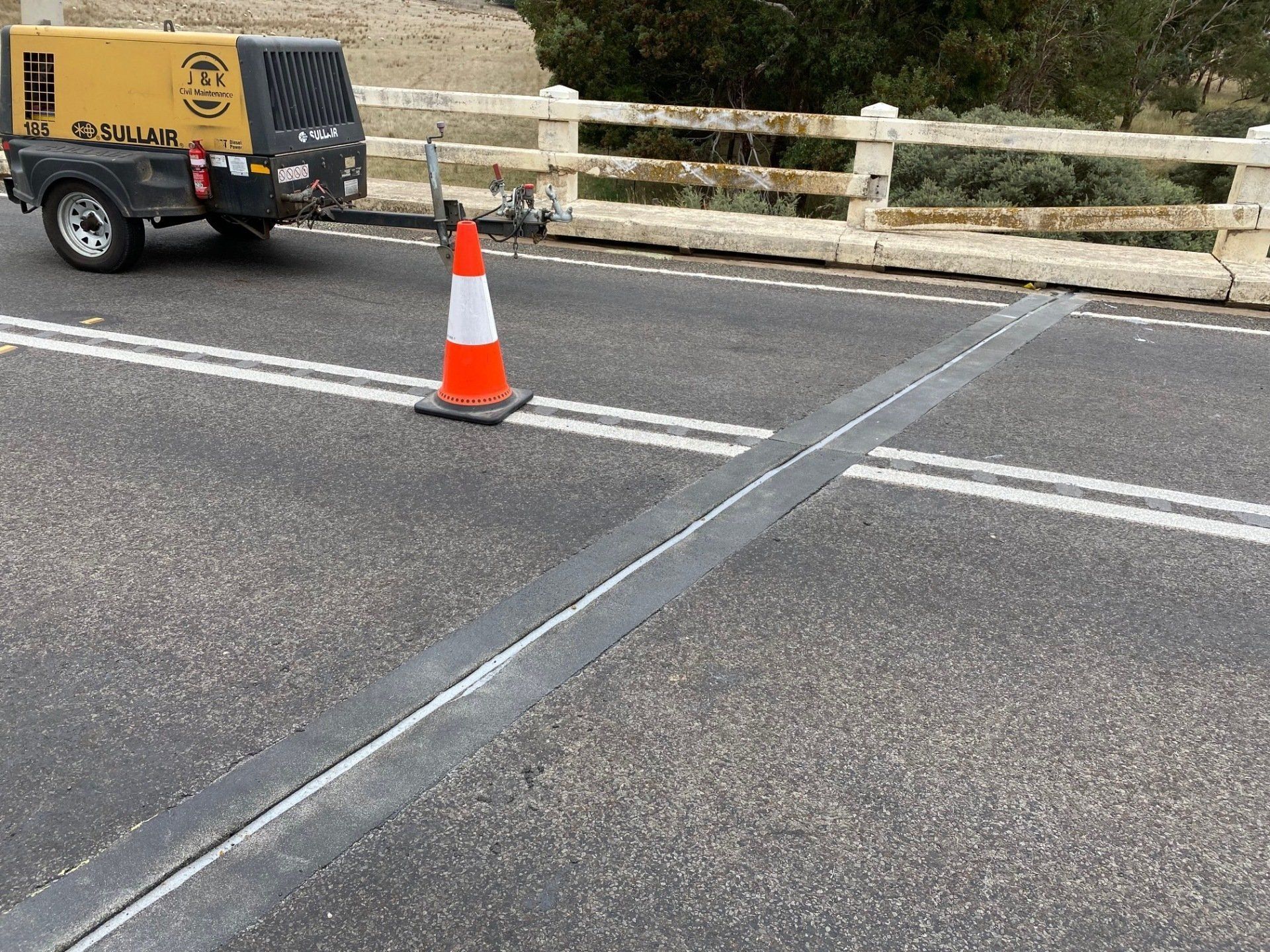 Orange traffic cone on a road near a yellow utility trailer and bridge railing.