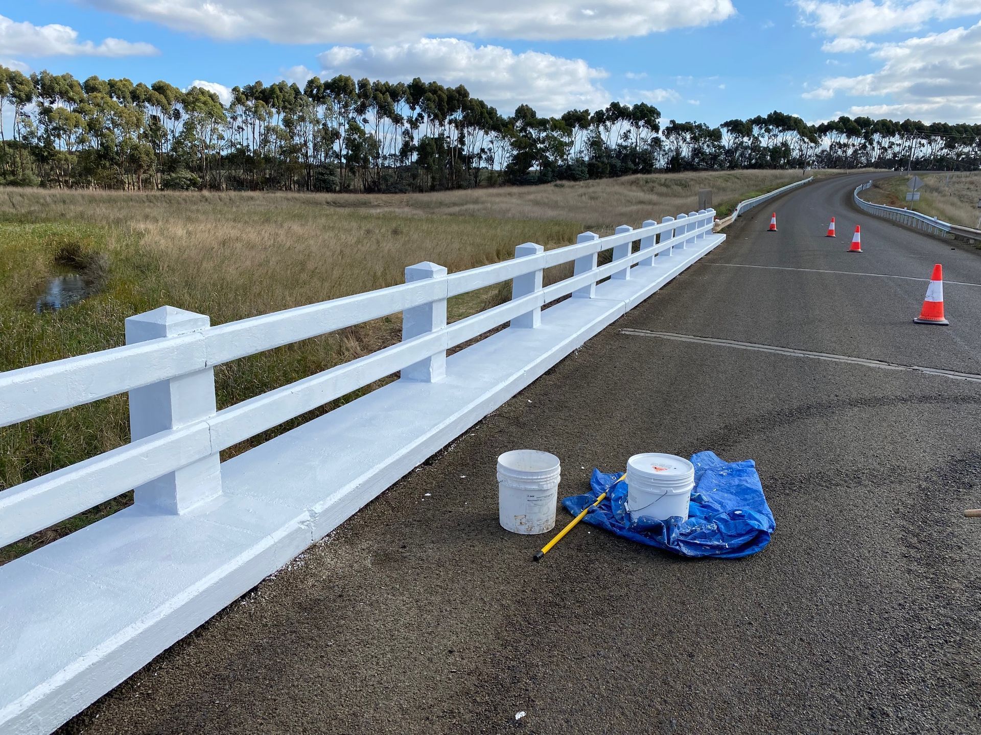 White roadside guardrail along a wet road with orange cones and paint supplies under a blue sky.