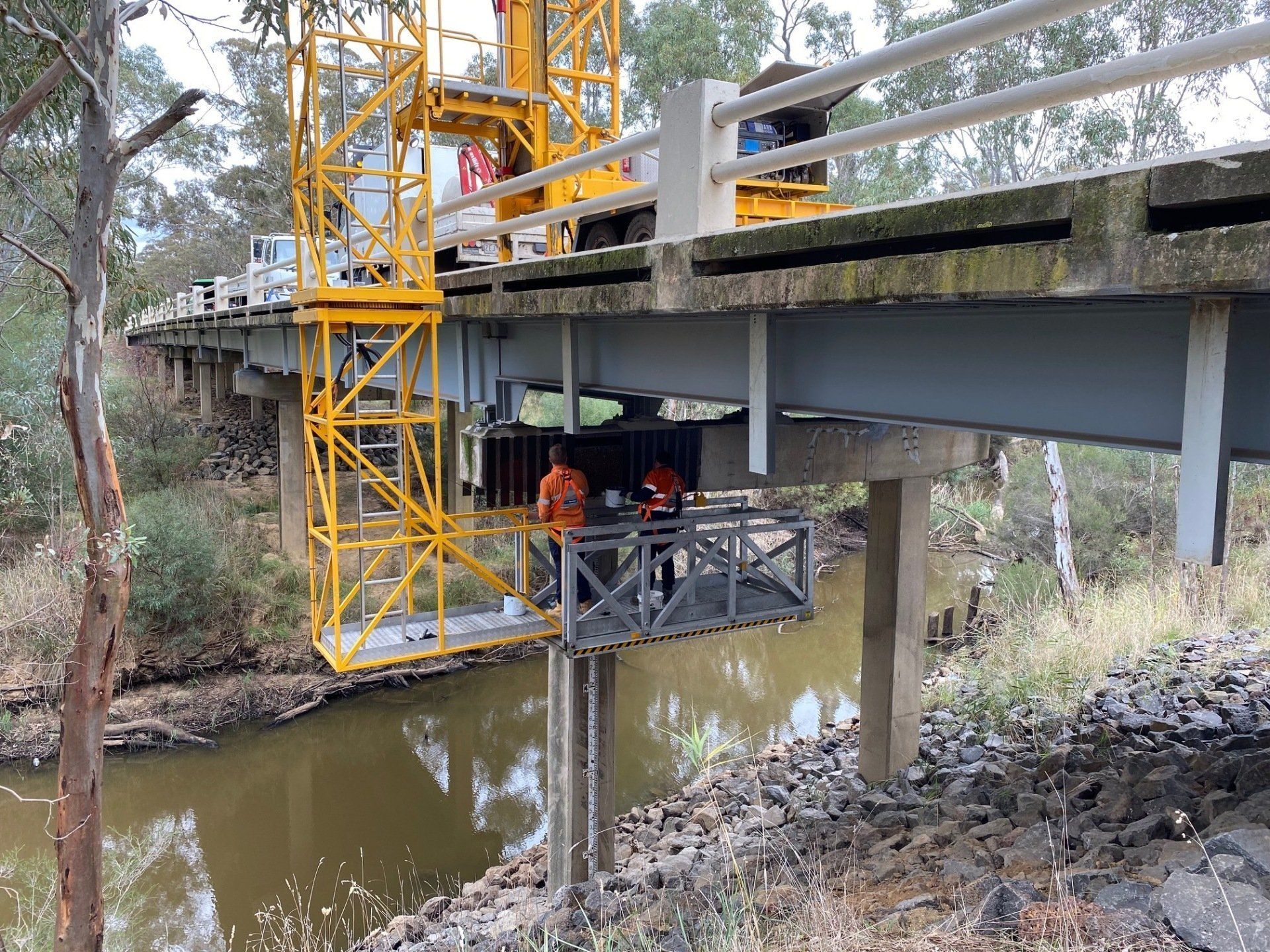 Workers on a bridge over a creek, with a yellow crane and a platform below.