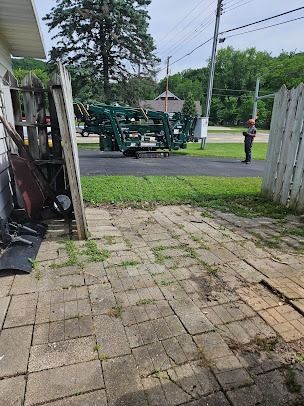 A green machine loading tree branches on a street, with a worker observing, viewed from a brick patio.