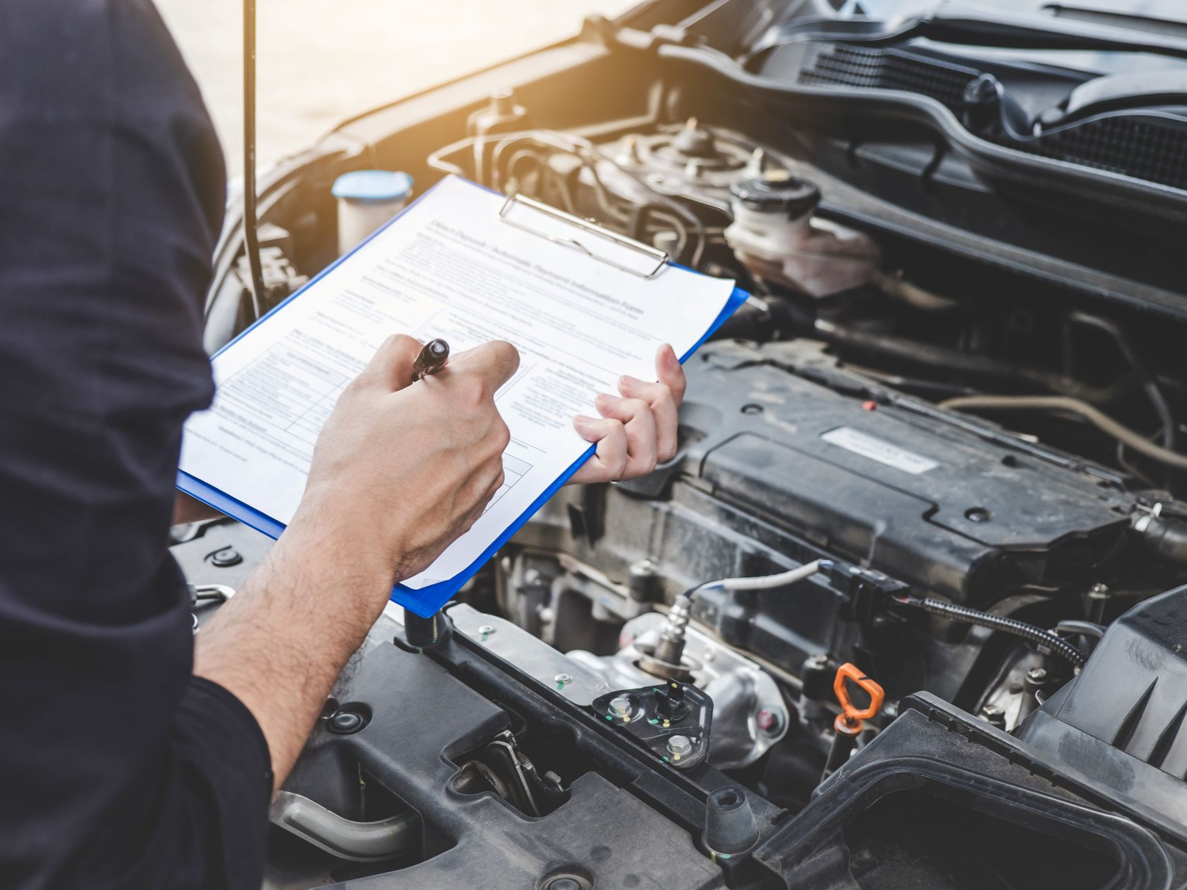 Mechanic filling out a clipboard checklist beside an open car engine in a repair shop