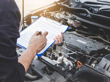 Mechanic writing on a clipboard beside an open car engine in a repair shop
